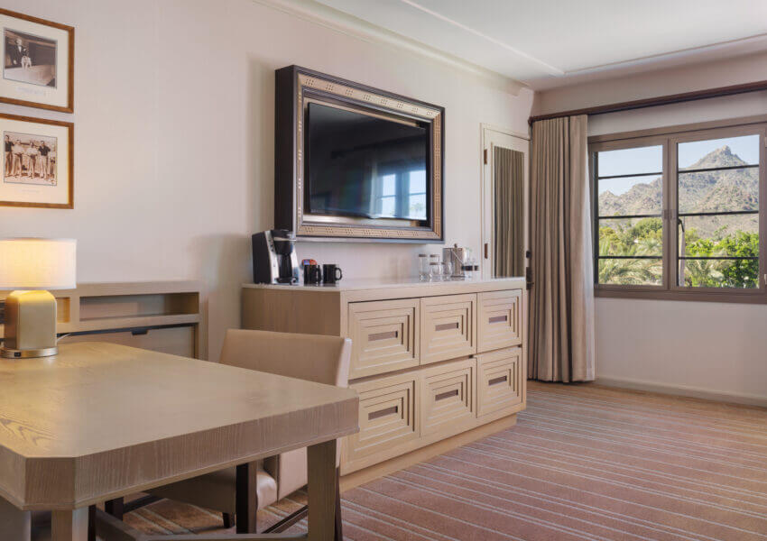 Hotel room with a desk, beige chair, lamp, wall-mounted TV, and dresser. Coffee maker and condiments are on the dresser. Two framed photos hang above. A window with curtains reveals a scenic mountain and greenery view. The room has a striped carpet flooring.