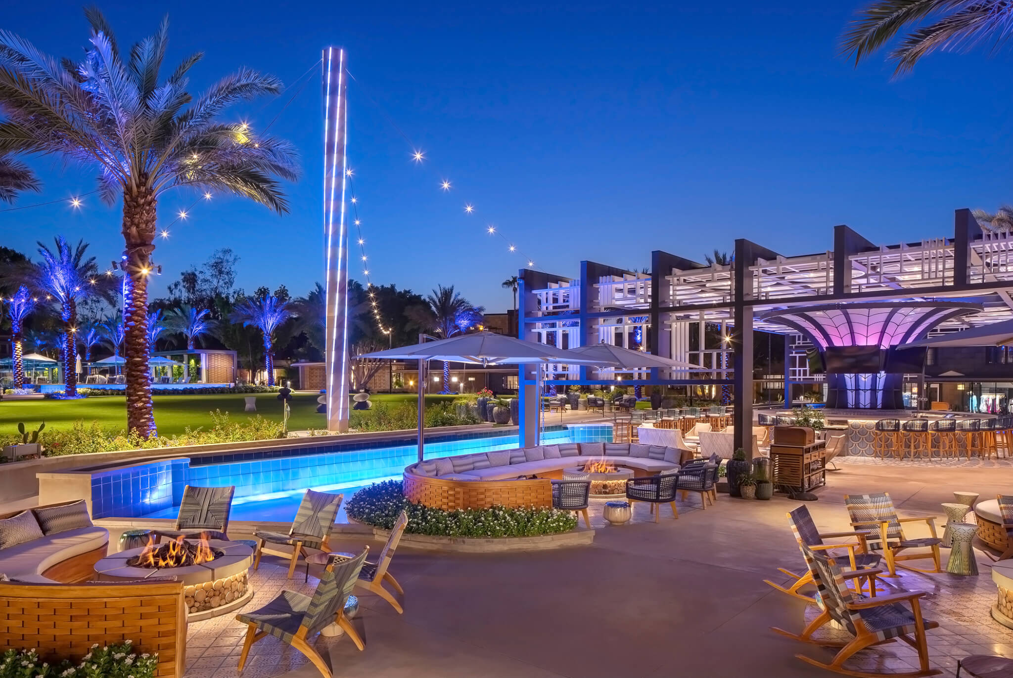A luxurious outdoor resort area at dusk, reminiscent of the Arizona Biltmore, featuring a pool surrounded by lounge chairs, umbrellas, and palm trees. String lights are draped across the space, creating a cozy ambiance. Fire pits and contemporary architecture complete the scene.