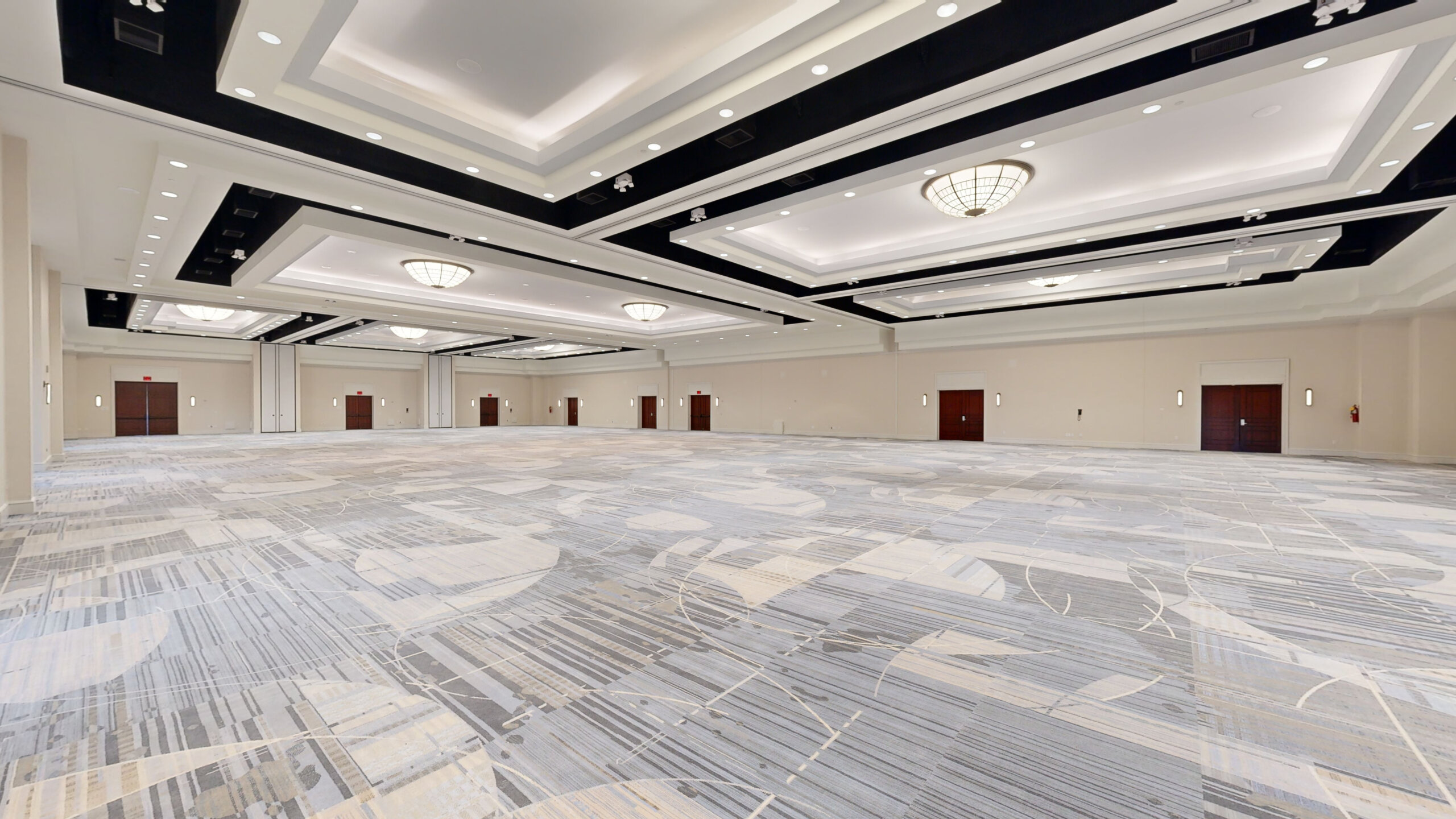 Spacious, empty ballroom with patterned carpet, high ceilings, modern chandeliers, and several closed wooden doors along the beige walls. Bright overhead lighting creates a clean, open atmosphere.