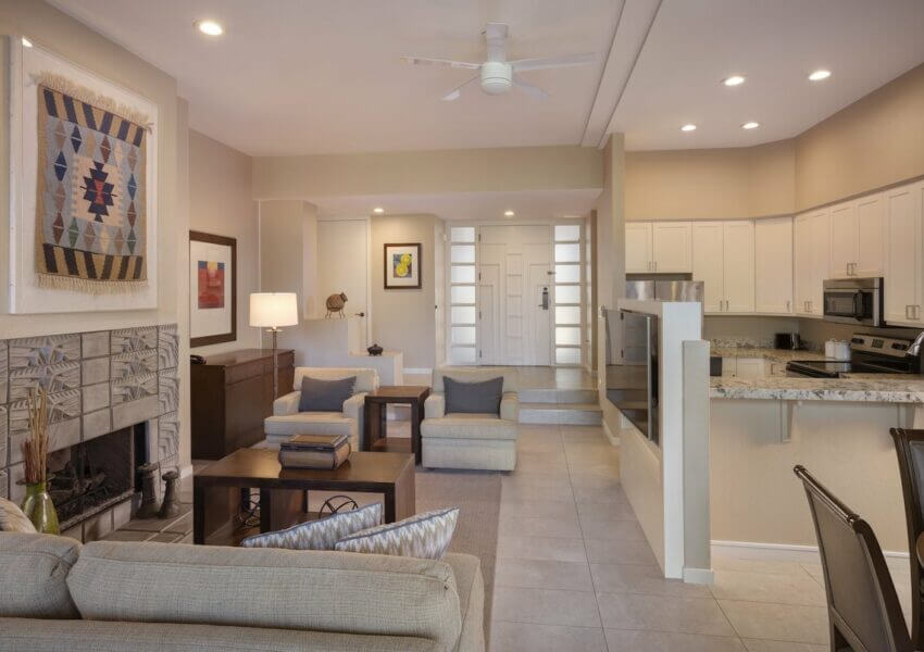 A modern living area with beige and white tones. Features a fireplace with tile design, wall art, and comfortable seating. Adjacent is a kitchen with white cabinets, stainless steel appliances, and a granite island. Recessed lighting and ceiling fan above.
