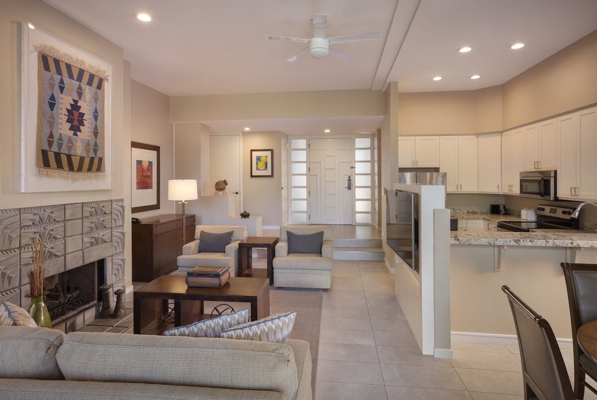 A modern living area with beige and white tones. Features a fireplace with tile design, wall art, and comfortable seating. Adjacent is a kitchen with white cabinets, stainless steel appliances, and a granite island. Recessed lighting and ceiling fan above.