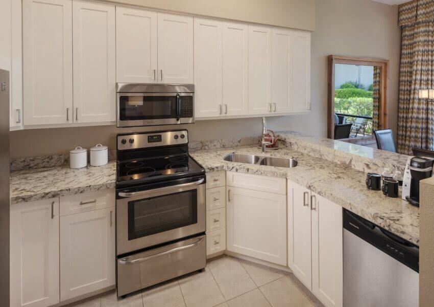 Modern kitchen with white cabinets and speckled granite countertops. Features stainless steel appliances, including an oven, microwave, and dishwasher. There's a double sink, canisters on the counter, and a coffee machine. A window shows a view of greenery outside.