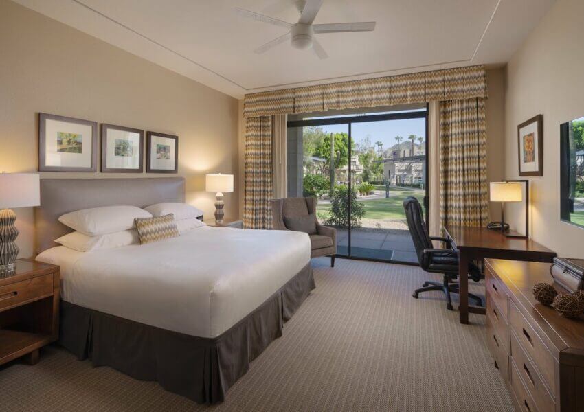 A spacious hotel room featuring a comfy queen-sized bed with white linens, wooden bedside tables with lamps, a plush grey armchair, a work desk with an office chair, and a large window with curtains providing a view of a patio and green outdoor scenery.