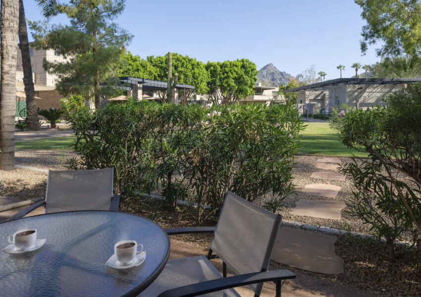 A serene outdoor patio features a round glass table with two chairs and two cups of coffee. The view includes lush greenery, palm trees, and manicured lawns leading to modern, sunlit buildings in the background, with a mountain visible under a clear blue sky.