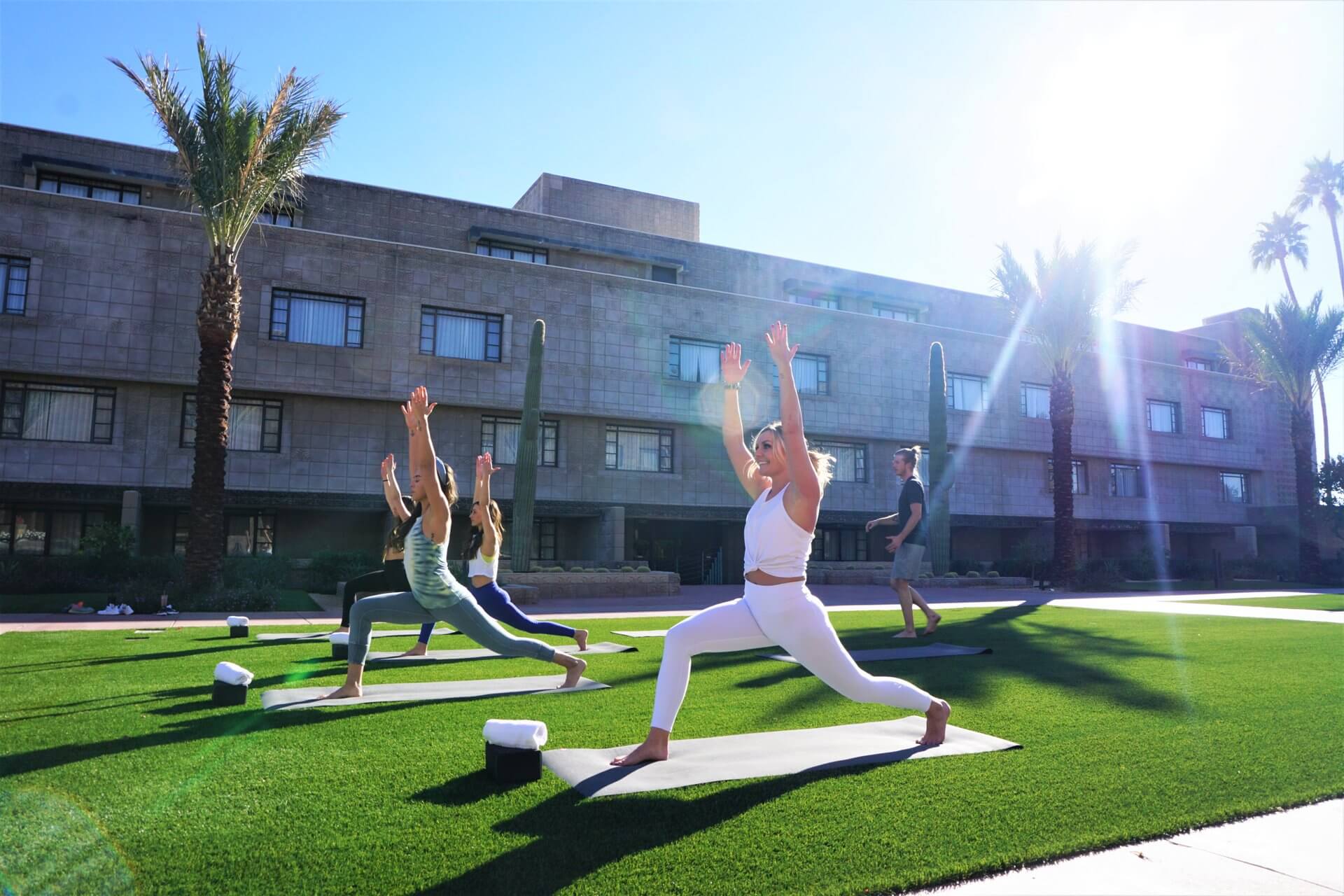 A group of people practice yoga outdoors on a sunny day, performing warrior poses on green grass. They are positioned in front of a large building with palm trees nearby. The sky is clear, and the sun casts a bright light over the scene.
